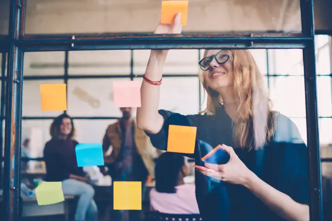 Lächeln junge Arbeitnehmerinnen in stilvollen Brillen mit Sticker für die Erstellungsstrategie des Arbeitsprojekts während der Brainstorming-Sitzung