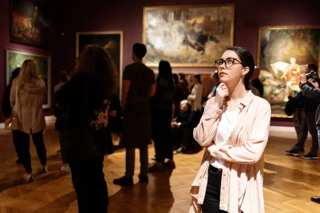 Achtsame junge kaukasische Frau, die Brille trägt und sich die Ausstellung ansieht. Im Hintergrund schauen die Leute Bilder an. Konzept des Museumstags